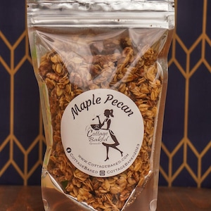 May include: A clear plastic bag filled with maple pecan granola. The bag has a white label with the text "Maple Pecan" and a logo for "Cottage Baked Granola". The label also includes the website address "www.cottagebaked.com" and social media handles for Facebook and Instagram.