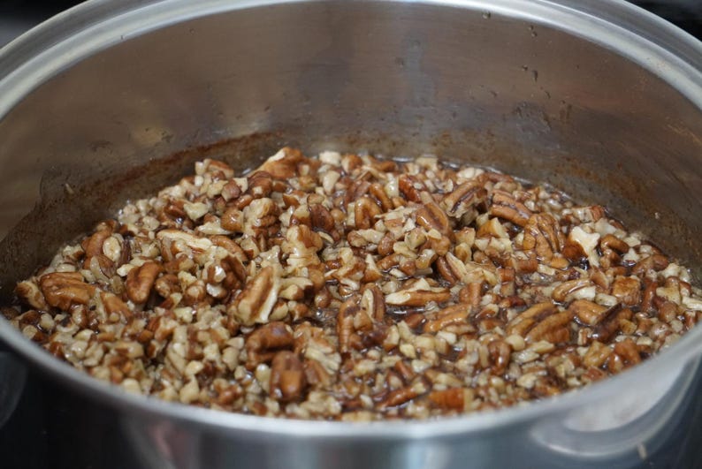 May include: A close-up of a pot filled with pecan pieces coated in a brown syrup. The pecans are evenly distributed throughout the pot.