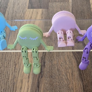 Op de afbeelding: Vier kleurrijke, grillige figuren in de vorm van macarons, met beweegbare ledematen en armen. De macarons zijn blauw, groen, roze en paars. De groene macaron heeft gesloten ogen, terwijl de paarse open ogen heeft. Ze worden tentoongesteld op een doorzichtige acryl plank.