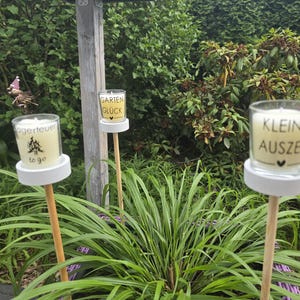 Pode incluir: Três velas de vidro em estacas de madeira num jardim. Cada vela tem texto, incluindo "Lagerfeuer to go", "Garten Glück" e "Kleine Auszeit". As velas estão sobre bases brancas, rodeadas por folhagem verde.