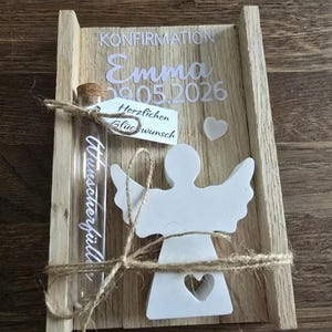 Könnte beinhalten: Eine Holzkiste mit dem Wort "Konfirmation" und dem Namen "Emma" mit dem Datum "09.05.2026" in weißer Schrift. Ein kleines Glasröhrchen mit Korken ist mit Bindfaden an der Kiste befestigt. Eine weiße Engelsfigur mit Herz-Ausschnitt ist ebenfalls angebracht.