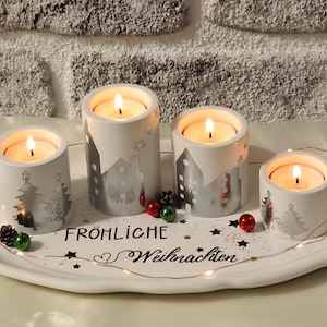 Op de afbeelding: Een feestelijke kerstkaarsen display. Vier witte kaarshouders met brandende kaarsen zijn gerangschikt op een wit ovaal dienblad. De houders hebben winterscène ontwerpen. De lade heeft de woorden "Fröhliche Weihnachten" en decoratieve lichten.