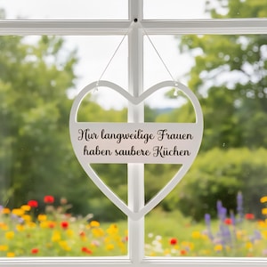 Könnte beinhalten: Ein weißes herzförmiges Holzschild, das vor einem Fenster hängt. Auf dem Schild steht in schwarzer Schrift der deutsche Text "Nur langweilige Frauen haben saubere Küchen". Der Hintergrund zeigt eine verschwommene Ansicht eines Gartens mit bunten Blumen.
