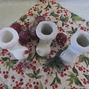 Könnte beinhalten: Drei weiße, stiefelförmige Keramikvasen sind auf einem Stoff mit floralem Muster mit roten Beeren und grünen Blättern angeordnet. Mehrere rote und burgunderfarbene dekorative Kugeln und ein kleines Bündel Kiefernnadeln sind ebenfalls vorhanden.