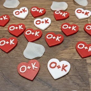 Può includere: Decorazioni a forma di cuore rosse e bianche con le lettere "O+K" in bianco. I cuori sono sparsi su una superficie di legno, con accenti bianchi a forma di petalo.