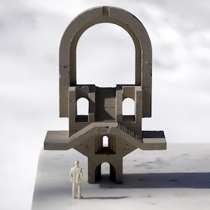 May include: A miniature architectural sculpture in light gray concrete, featuring arches, staircases, and geometric forms. A small white figurine stands in front of the structure, placed on a white surface with a snowy background.