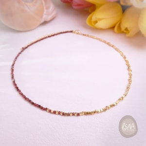 May include: A delicate necklace featuring a half-and-half design. One side is adorned with small, deep red beads, while the other side showcases gold-colored, geometric beads. The necklace is displayed on a white surface with yellow tulips in the background.