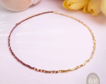May include: A delicate necklace featuring a half-and-half design. One side is adorned with small, deep red beads, while the other side showcases gold-colored, geometric beads. The necklace is displayed on a white surface with yellow tulips in the background.