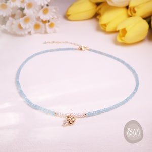 May include: A light blue beaded necklace featuring a gold-toned heart charm with a clear teardrop crystal. The necklace includes a section of small white pearls near the center and a gold-toned metal clasp. The text RENA DESIGN is visible.