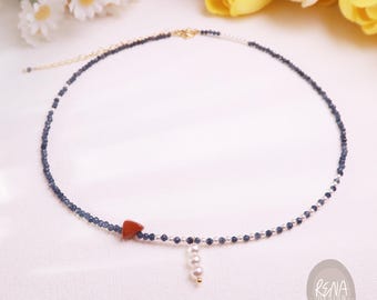 Op de afbeelding: Een kralenketting met kleine donkerblauwe gefacetteerde kralen, een enkele driehoekige rode steen en een verticale hanger bestaande uit drie kleine witte parels. De ketting is voorzien van een goudkleurige sluiting en een verlengketting.