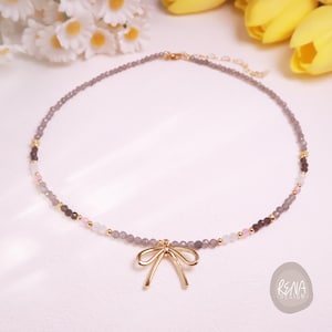 Peut inclure: Un collier d&eacute;licat compos&eacute; de petites perles grises, avec des touches de rose, de blanc et de marron fonc&eacute;. Un pendentif en forme de n&oelig;ud dor&eacute; est suspendu au centre. Le collier est pr&eacute;sent&eacute; sur une surface blanche, avec des fleurs en arri&egrave;re-plan.
