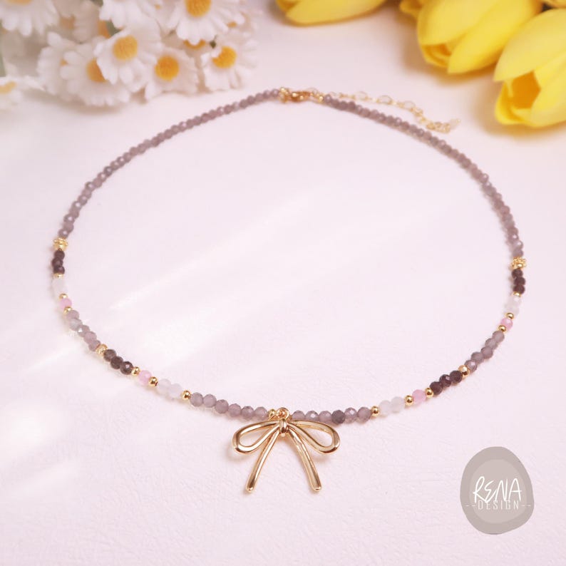 Peut inclure: Un collier d&eacute;licat compos&eacute; de petites perles grises, avec des touches de rose, de blanc et de marron fonc&eacute;. Un pendentif en forme de n&oelig;ud dor&eacute; est suspendu au centre. Le collier est pr&eacute;sent&eacute; sur une surface blanche, avec des fleurs en arri&egrave;re-plan.