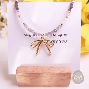 Peut inclure: Un collier d&eacute;licat avec un pendentif en forme de n&oelig;ud dor&eacute; et une cha&icirc;ne perl&eacute;e avec des accents gris, roses et dor&eacute;s. Le collier est pr&eacute;sent&eacute; sur une carte blanche avec le texte "May the rise up to MEET YOU".