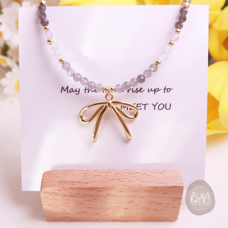 Peut inclure: Un collier d&eacute;licat avec un pendentif en forme de n&oelig;ud dor&eacute; et une cha&icirc;ne perl&eacute;e avec des accents gris, roses et dor&eacute;s. Le collier est pr&eacute;sent&eacute; sur une carte blanche avec le texte "May the rise up to MEET YOU".