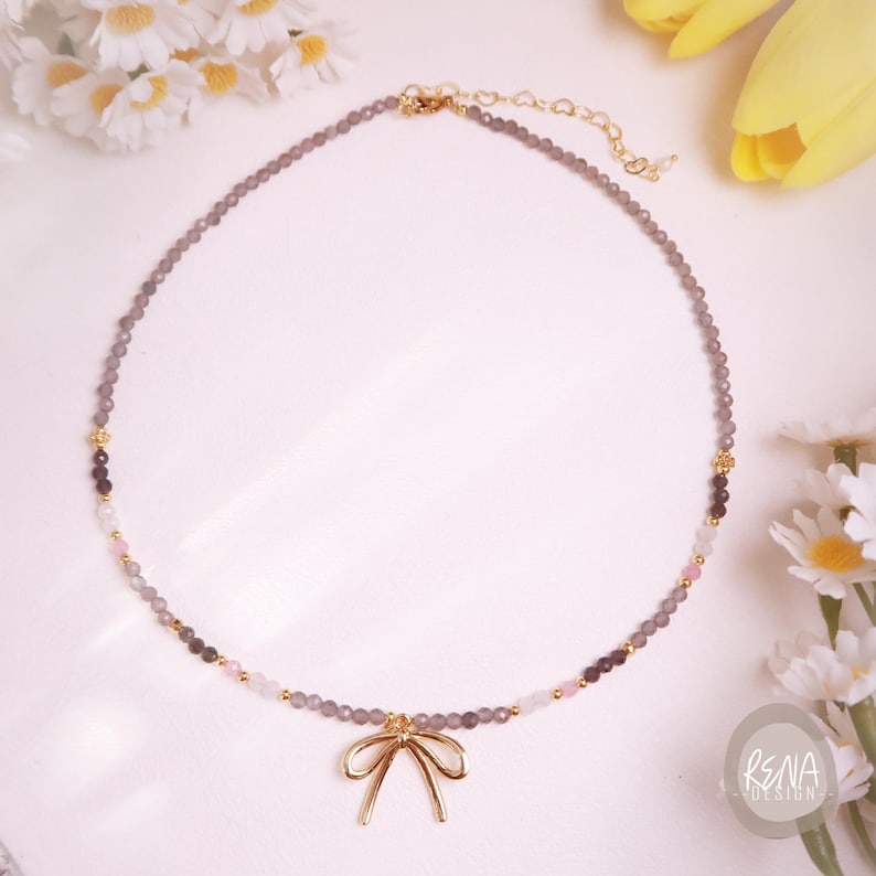 Peut inclure: Un collier d&eacute;licat compos&eacute; d'un fil de petites perles grises avec des accents de perles roses, blanches et gris fonc&eacute;. Un pendentif en forme de n&oelig;ud dor&eacute; est suspendu au centre. Le collier est pr&eacute;sent&eacute; sur une surface blanche avec des fleurs.