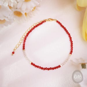 May include: A delicate red beaded bracelet with gold-colored chain and accents. The bracelet features small, faceted red beads and white pearl-like beads. The adjustable chain adds versatility to the design. The bracelet is a stylish accessory.
