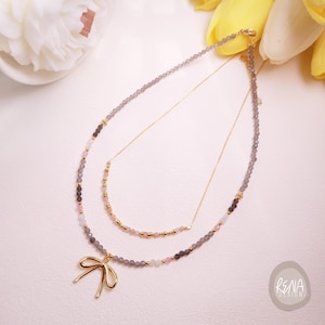 Peut inclure: Un collier &agrave; plusieurs rangs avec un pendentif en forme de n&oelig;ud dor&eacute;. Le collier est orn&eacute; de perles grises, roses et dor&eacute;es. La couche sup&eacute;rieure est une fine cha&icirc;ne dor&eacute;e.