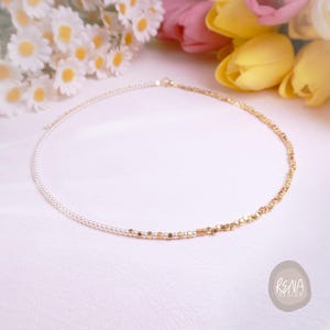 May include: A delicate necklace featuring a half-and-half design. One side is composed of small, white pearls, while the other is adorned with gold-colored, geometric beads. The necklace is displayed on a white surface with flowers in the background.
