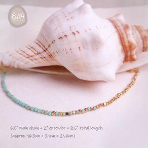 May include: A delicate gold-toned necklace with turquoise beads and gold-colored square beads. The main chain is 6.5 inches, with a 2-inch extender, totaling 8.5 inches. The necklace is displayed near a large seashell.