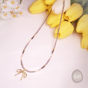 Peut inclure: Un collier d&eacute;licat compos&eacute; de perles grises et roses, agr&eacute;ment&eacute; d'entretoises dor&eacute;es. Le collier est orn&eacute; d'un pendentif en forme de n&oelig;ud dor&eacute;. Le fond comprend des tulipes jaunes et des fleurs blanches.