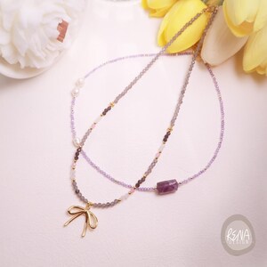 Peut inclure: Deux colliers d&eacute;licats. L'un pr&eacute;sente un pendentif en forme de n&oelig;ud dor&eacute; et des perles grises, l'autre une pierre pr&eacute;cieuse violette et des perles lavande. Les deux colliers ont des accents dor&eacute;s et sont pr&eacute;sent&eacute;s sur une surface blanche.