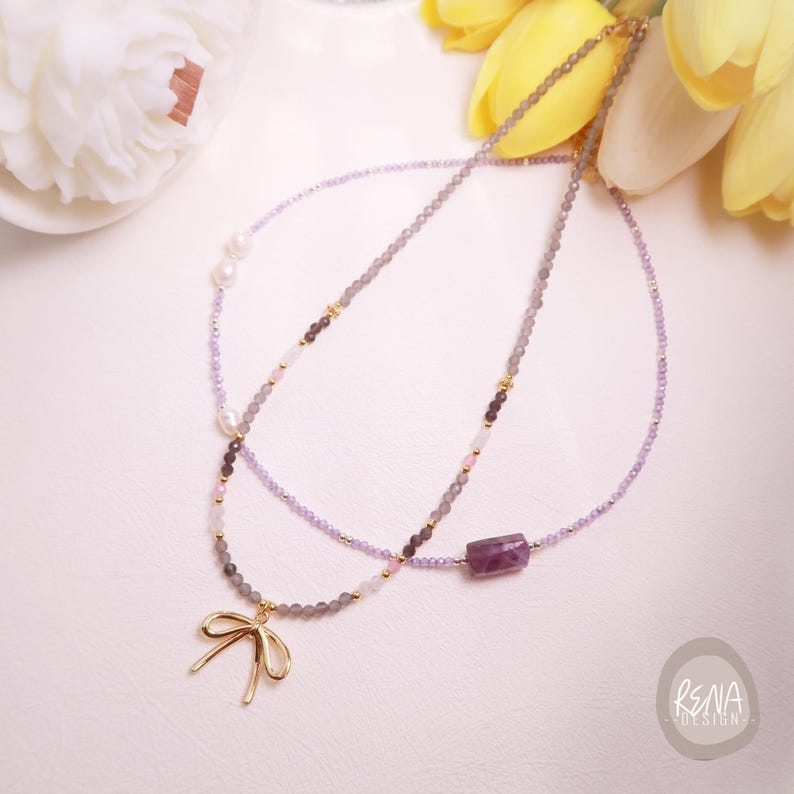 Peut inclure: Deux colliers d&eacute;licats. L'un pr&eacute;sente un pendentif en forme de n&oelig;ud dor&eacute; et des perles grises, l'autre une pierre pr&eacute;cieuse violette et des perles lavande. Les deux colliers ont des accents dor&eacute;s et sont pr&eacute;sent&eacute;s sur une surface blanche.