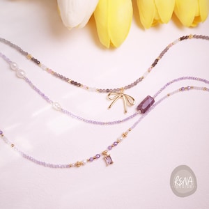 Peut inclure: Trois colliers d&eacute;licats avec divers breloques et perles. L'un des colliers pr&eacute;sente un pendentif en forme de n&oelig;ud dor&eacute;, un autre une gemme rectangulaire violette, et le troisi&egrave;me un petit pendentif en forme de gemme violette. Les colliers sont de tons violets, gris et dor&eacute;s.