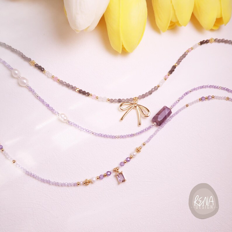 Peut inclure: Trois colliers d&eacute;licats avec divers breloques et perles. L'un des colliers pr&eacute;sente un pendentif en forme de n&oelig;ud dor&eacute;, un autre une gemme rectangulaire violette, et le troisi&egrave;me un petit pendentif en forme de gemme violette. Les colliers sont de tons violets, gris et dor&eacute;s.