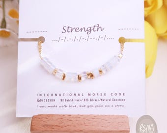 Puede incluir: Una pulsera chapada en oro con piedras preciosas naturales y detalles dorados. La pulsera se muestra en una tarjeta blanca con la palabra "Strength" y código Morse. La tarjeta también incluye el texto "International Morse Code".
