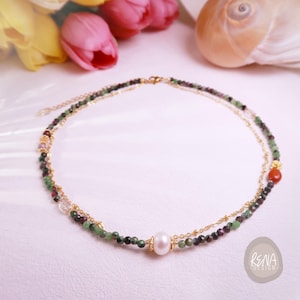 May include: A delicate, layered necklace featuring a central pearl and a mix of green, red, and clear beads. The necklace has a gold chain and clasp. A small, round, orange bead is visible on the lower layer. The necklace is set against a soft, light background.
