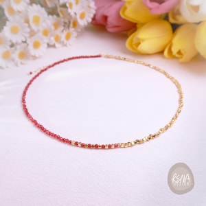 May include: A delicate necklace featuring a half-and-half design. One side is composed of small, vibrant red beads, while the other side is made of gold-colored, faceted beads. The necklace is displayed on a white surface with flowers in the background.