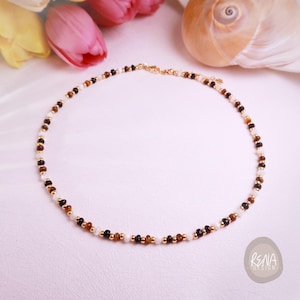 Puede incluir: Delicado collar de cuentas con una mezcla de cuentas blancas, marrones y negras, intercaladas con pequeñas cuentas doradas. El collar se exhibe sobre una superficie blanca, con flores rosas y amarillas en el fondo.