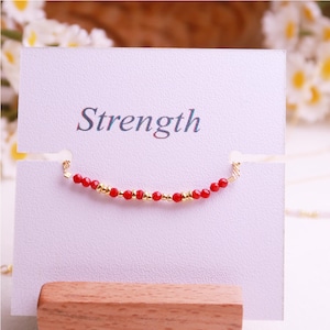 May include: A delicate bracelet featuring small red faceted beads and gold-toned metallic spacers, displayed on a white card with the word Strength printed in blue text. The jewelry piece is secured through two horizontal slits in the card.