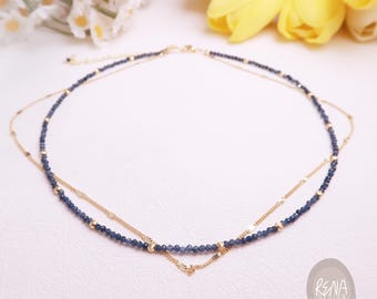 Könnte beinhalten: Eine mehrlagige Halskette mit einer Reihe kleiner, facettierter dunkelblauer Perlen, die mit winzigen goldfarbenen Metallkugeln und einer zarten goldfarbenen Kette kombiniert sind. Das Schmuckstück enthält ein kleines, rundes Logo-Etikett mit dem Text RENA DESIGN.