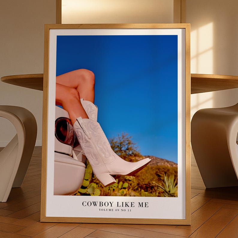 May include: A framed print featuring a pair of white cowboy boots with a pointed toe, set against a blue sky and desert landscape. The text "COWBOY LIKE ME" and "VOLUME 09 NO 11" are at the bottom of the print.