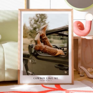 May include: A framed print featuring brown cowboy boots extending from a dark-colored vehicle. The boots have decorative stitching. The print includes the text "COWBOY LIKE ME" and "VOLUME 09 NO 11" at the bottom. The frame is light-colored.