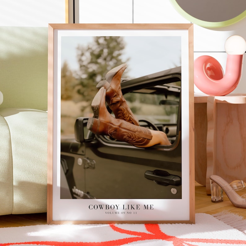 May include: A framed print featuring brown cowboy boots extending from a dark-colored vehicle. The boots have decorative stitching. The print includes the text "COWBOY LIKE ME" and "VOLUME 09 NO 11" at the bottom. The frame is light-colored.