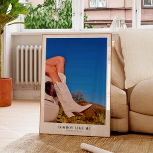 May include: A framed art print featuring a photograph of a person wearing white cowboy boots. The boots are set against a blue sky and desert landscape. The text "COWBOY LIKE ME" is printed at the bottom.