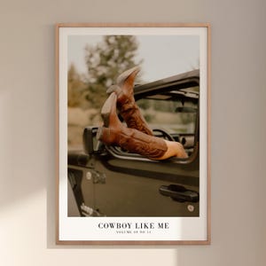 May include: A framed print featuring brown cowboy boots extending from a dark-colored vehicle. The boots have decorative stitching. The image has the text "COWBOY LIKE ME" and "VOLUME 09 NO 11" at the bottom.
