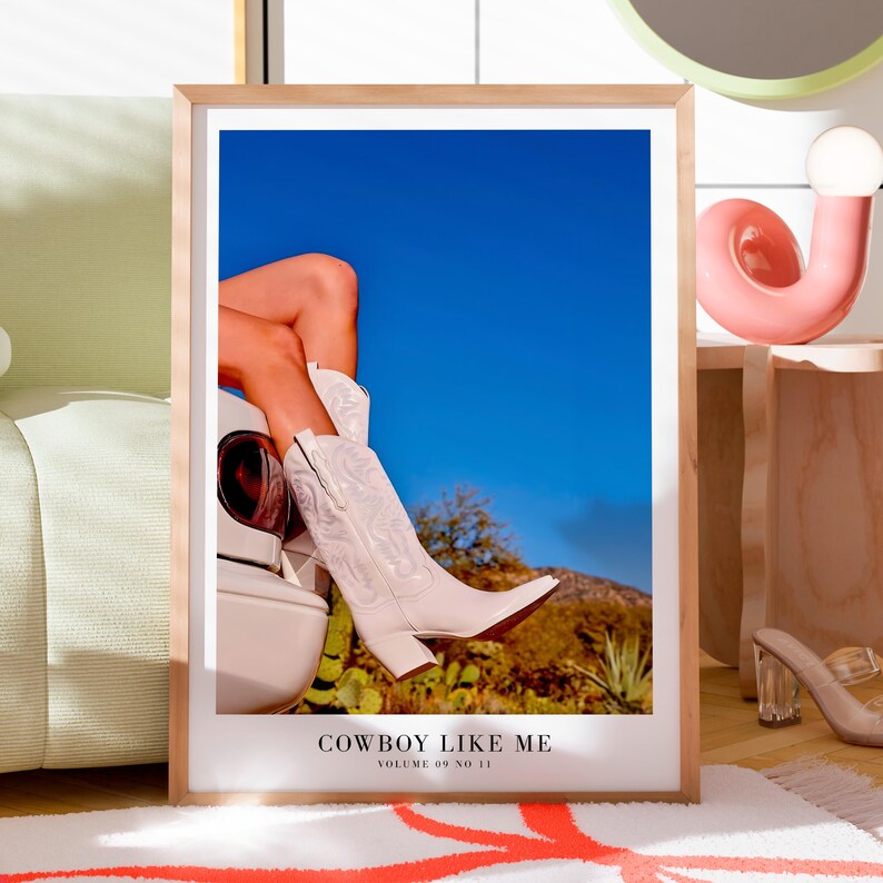 May include: A framed print featuring white cowboy boots with intricate stitching, set against a backdrop of a blue sky and desert landscape. The text "COWBOY LIKE ME" and "VOLUME 09 NO 11" are at the bottom of the print.