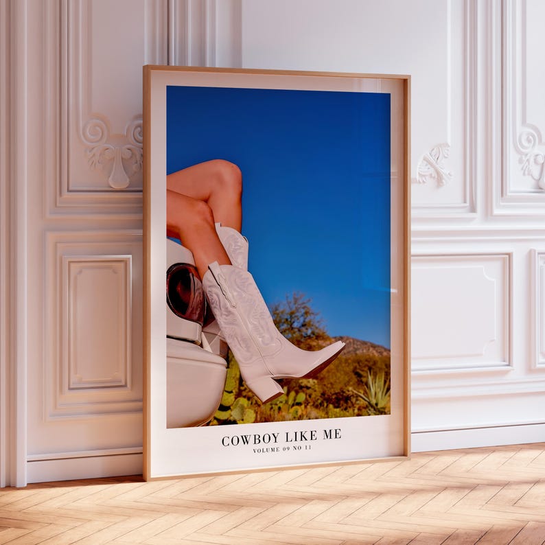 May include: A framed print featuring a pair of white cowboy boots with intricate stitching, set against a backdrop of a clear blue sky and desert landscape. The print includes the text "COWBOY LIKE ME" and "VOLUME 09 NO 11".