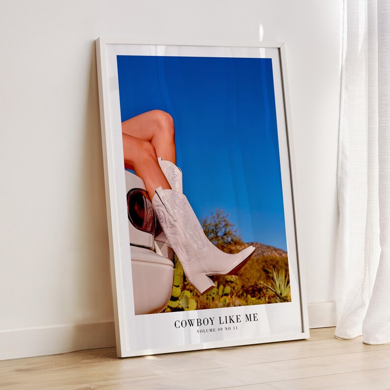 May include: A framed print featuring a pair of white cowboy boots. The boots are worn by a person sitting on a car, with a blue sky and desert landscape in the background. The text "COWBOY LIKE ME" is at the bottom.