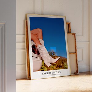 May include: A framed art print featuring a photograph of white cowboy boots and legs against a blue sky and desert landscape. The text "COWBOY LIKE ME" is visible at the bottom of the print.