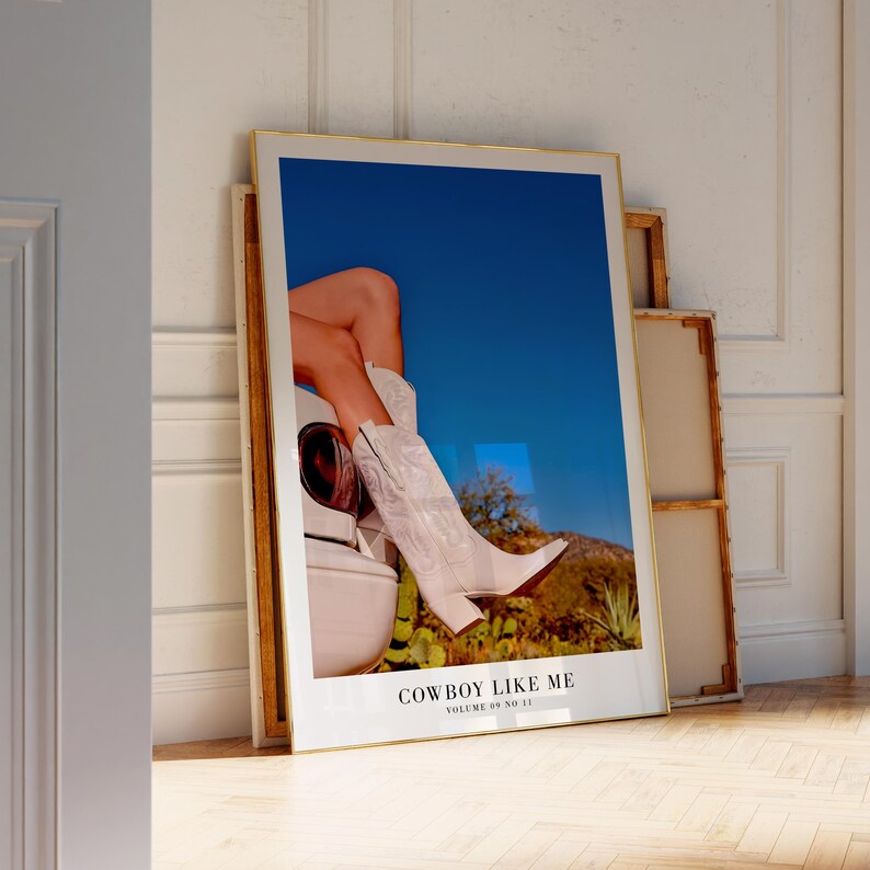 May include: A framed art print featuring a photograph of white cowboy boots and legs against a blue sky and desert landscape. The text "COWBOY LIKE ME" is visible at the bottom of the print.