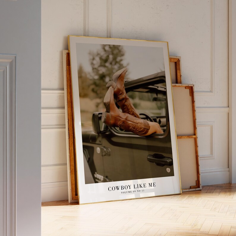 May include: A framed print featuring a photograph of brown cowboy boots extending from a dark green vehicle. The text "COWBOY LIKE ME" is visible at the bottom of the print. The print is leaning against a white wall.