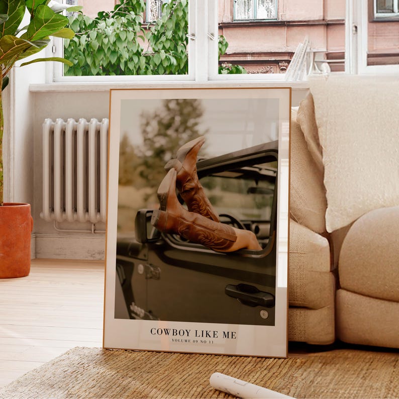 May include: Framed print featuring brown cowboy boots with decorative stitching, resting on the dashboard of a dark-colored vehicle. The print includes the text "COWBOY LIKE ME" and "VOLUME 89 NO 11". The image is set against a blurred outdoor backdrop.