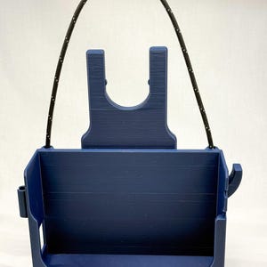May include: A navy blue, 3D-printed carrying case with a handle made of black and white speckled cord. The case has a rectangular base and a vertical support with a curved top. The handle is attached to the top of the support.