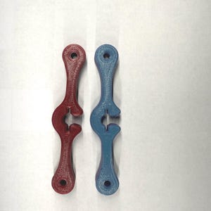 Könnte beinhalten: Zwei 3D-gedruckte Werkzeuge, eines rot und eines blau, werden vor einem weißen Hintergrund gezeigt. Jedes Werkzeug hat eine einzigartige, gebogene Form mit einem Loch oben und einer gebogenen Öffnung unten. Die Werkzeuge scheinen aus Kunststoff zu sein.