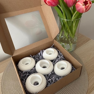 Op de afbeelding: Een set van vijf witte keramische kaarsenhouders met een geribbeld ontwerp, verpakt in een bruine doos met een transparant venster. De doos is gevuld met gescheurd papier en de kaarsenhouders zijn in een cirkel gerangschikt. Op de achtergrond staat een boeket roze tulpen.