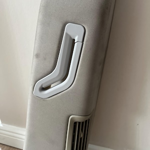 Op de afbeelding: Een lichtgrijs auto-interieurpaneel met een ingebouwde handgreep en ventilatierooster. De handgreep is van gebogen, gebroken wit plastic en het rooster heeft verticale lamellen. Het paneel heeft een textieloppervlak.